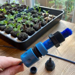 Close-up of a blue irrigation connector being assembled next to a seedling tray with young plants.